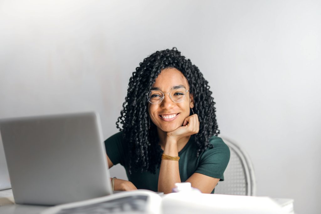 Joyful businesswoman with curly hair smiling at camera while using laptop indoors.